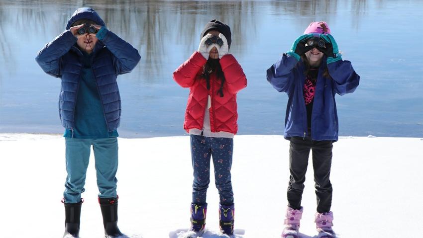 Three kids holding binoculars explore Aurora's wintery backdrop.